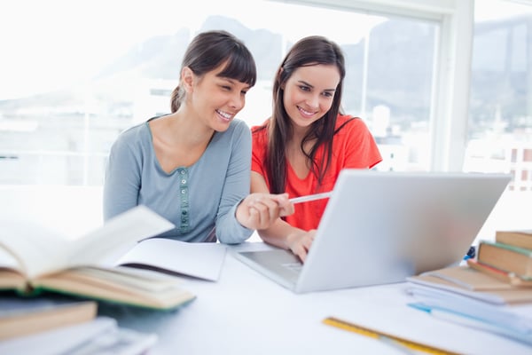 The girls are both sitting beside each other and smiling as they use the laptop as one girl points to something on screen-1